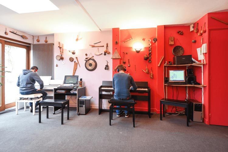 Deux personnes jouent du clavier dans une salle de musique dont les murs sont décorés d'instruments. L'une d'elles utilise un ordinateur.