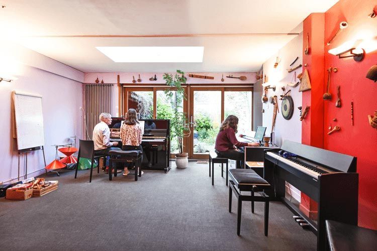Trois personnes dans un studio de musique, deux aux pianos, une à un bureau. Des instruments sont exposés sur un mur rouge.
