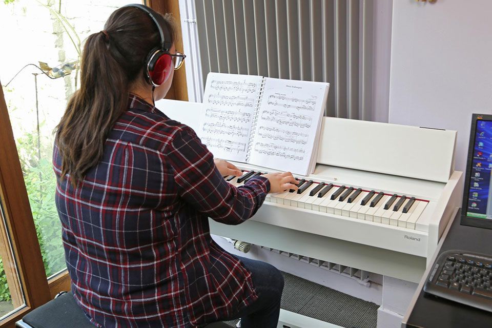 Une personne portant un casque audio joue du piano blanc avec des partitions ; près d'une fenêtre et d'un ordinateur.