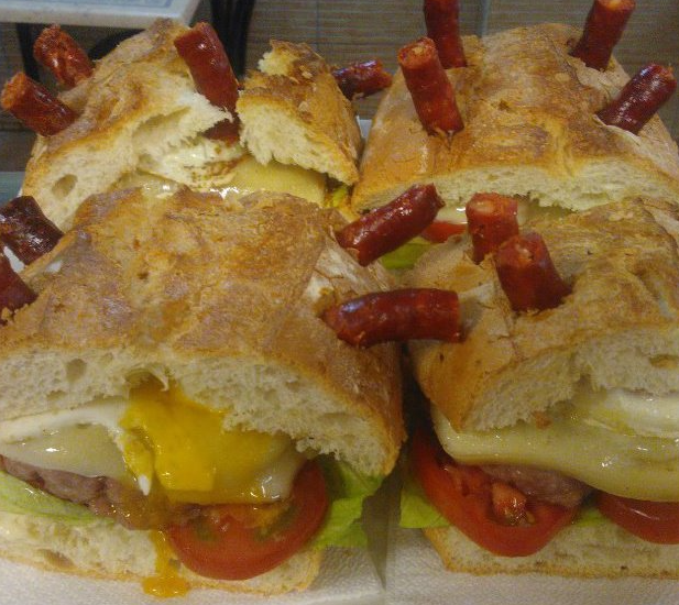Dos bocadillos en pan crujiente, cubiertos con salchichas peque&ntilde;as y rellenos de huevo frito, queso, lechuga y tomate.