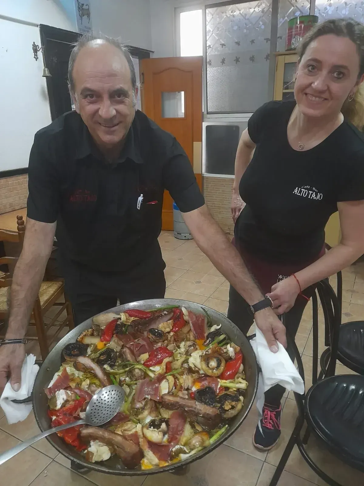 Un hombre y una mujer en un restaurante sostienen una gran paellera y sonríen a la cámara.