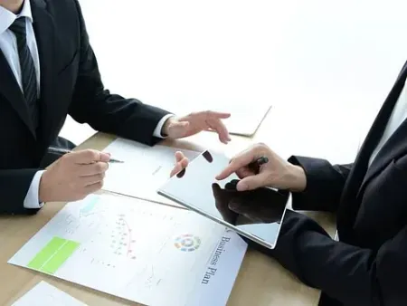 Dos personas de traje sentadas en un escritorio, revisando documentos y una tableta. Una de ellas señala con un bolígrafo.