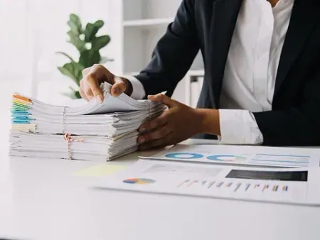 Una persona con traje revisa una gran pila de documentos en un escritorio con gráficos y una planta en el fondo.