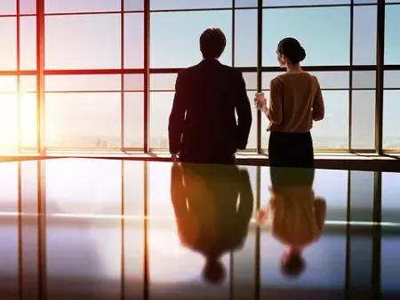 Dos hombres de negocios observan desde grandes ventanales, con reflejos visibles en el suelo pulido. El sol brilla con fuerza.