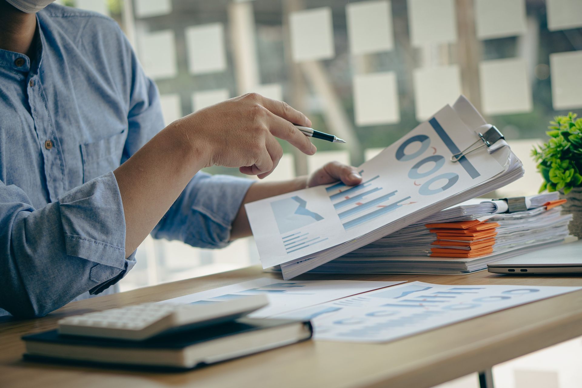 Hombre con camisa azul señalando gráficos financieros con un bolígrafo, trabajando en un escritorio con calculadora, cuaderno y papeleo.