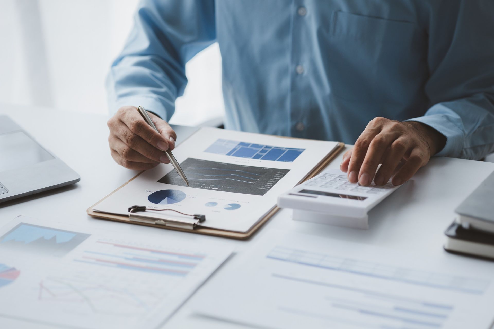 Persona con una camisa azul analizando datos financieros con una calculadora y un bolígrafo en un escritorio, rodeada de gráficos y documentos.