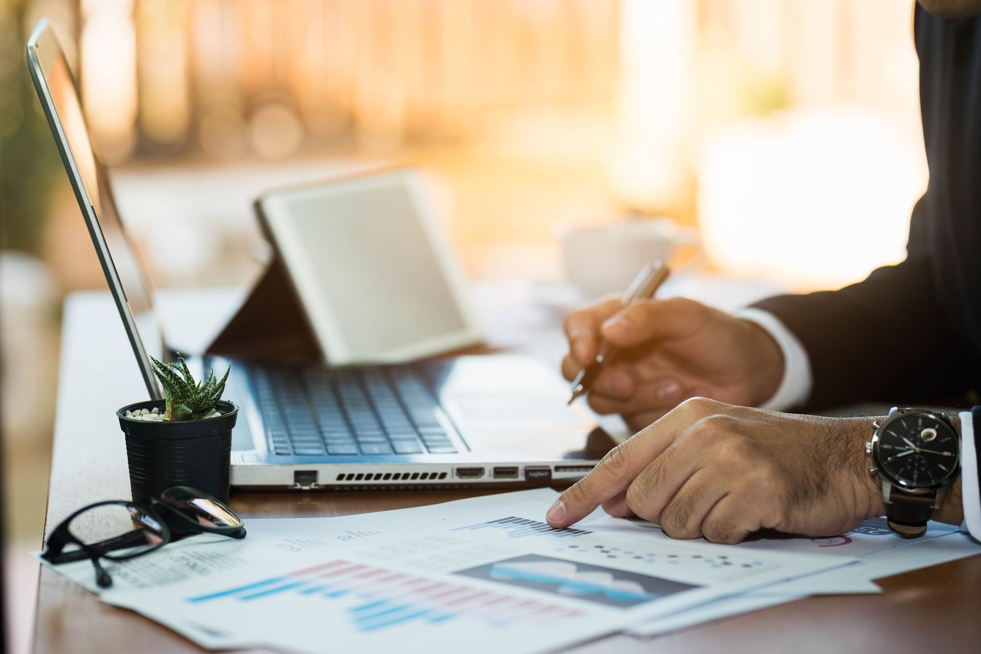 Un hombre con traje analiza datos financieros en un escritorio, señalando un gráfico con un bolígrafo; también hay una computadora portátil, gafas y una planta.