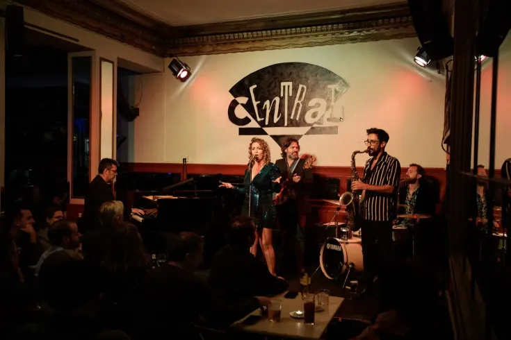 Banda de jazz en vivo tocando en un club con poca luz: cantante, saxofonista, pianista, baterista.