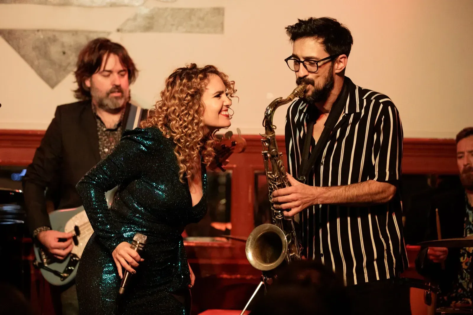 Banda tocando en vivo: cantante con vestido de lentejuelas, saxofonista, guitarrista y baterista en un lugar poco iluminado.