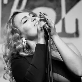Mujer cantando frente a un micrófono, con los ojos cerrados y cabello rubio y rizado. Foto en blanco y negro.