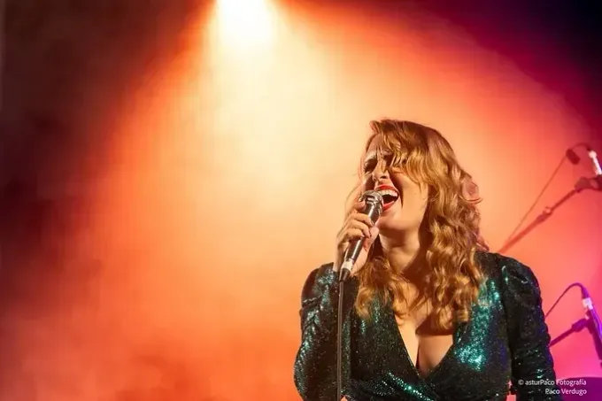Mujer cantando frente a un micrófono en el escenario bajo una iluminación naranja, vistiendo un vestido verde brillante.