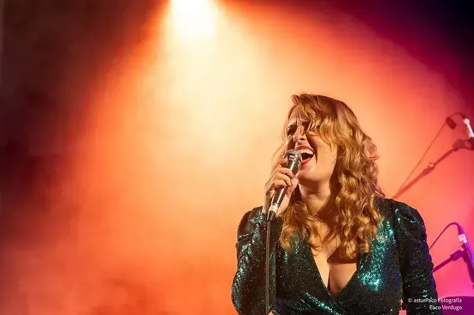 Mujer cantando en el escenario con un top verde de lentejuelas. Iluminación naranja.