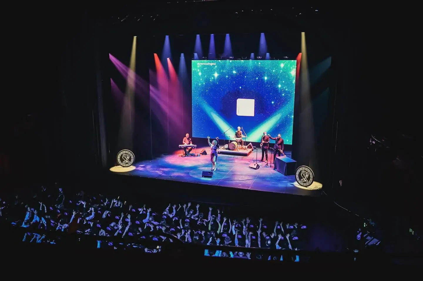 Banda actuando en un escenario con luces de colores y una pantalla de fondo, frente a un público que la anima.