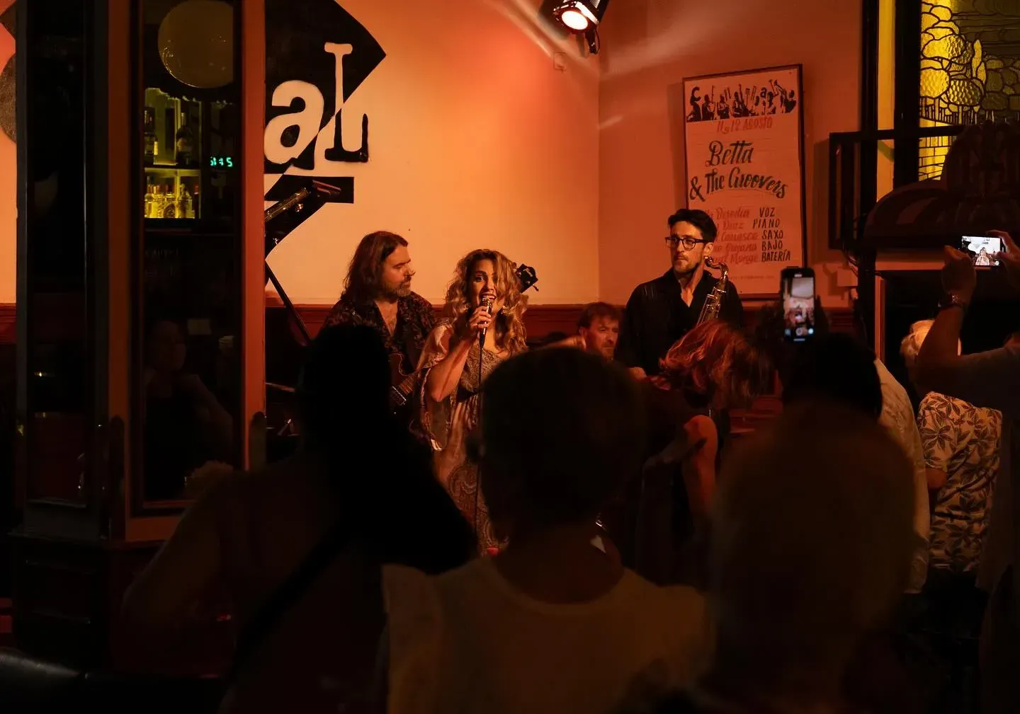 Banda tocando en vivo en un lugar poco iluminado mientras el público observa y filma con sus teléfonos.