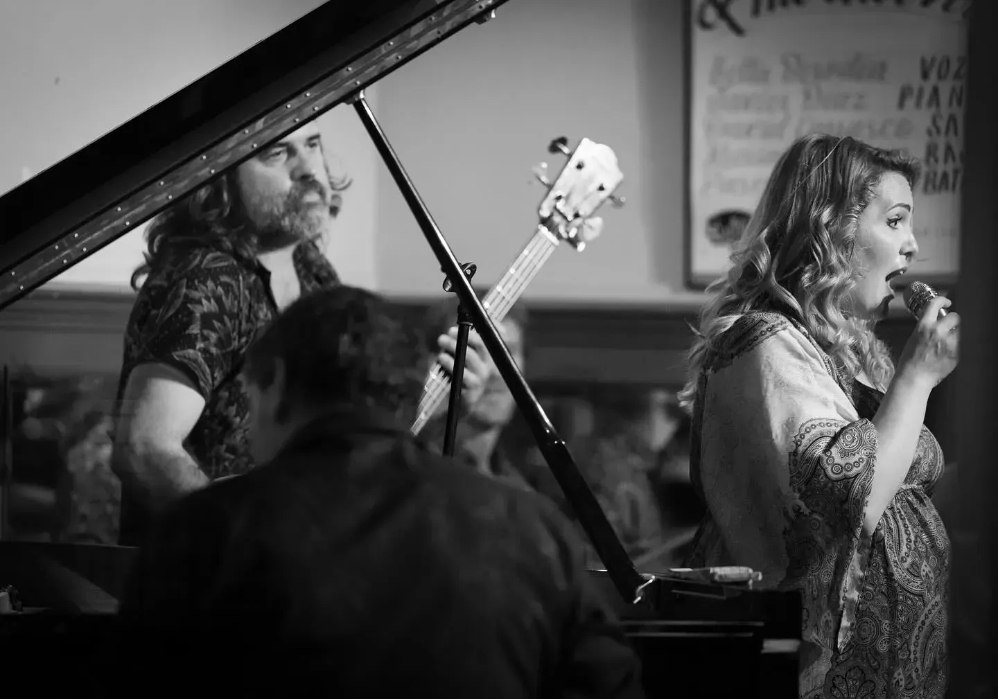 Una actuación de jazz: una mujer canta, un bajista y un pianista al piano en un lugar con poca luz.