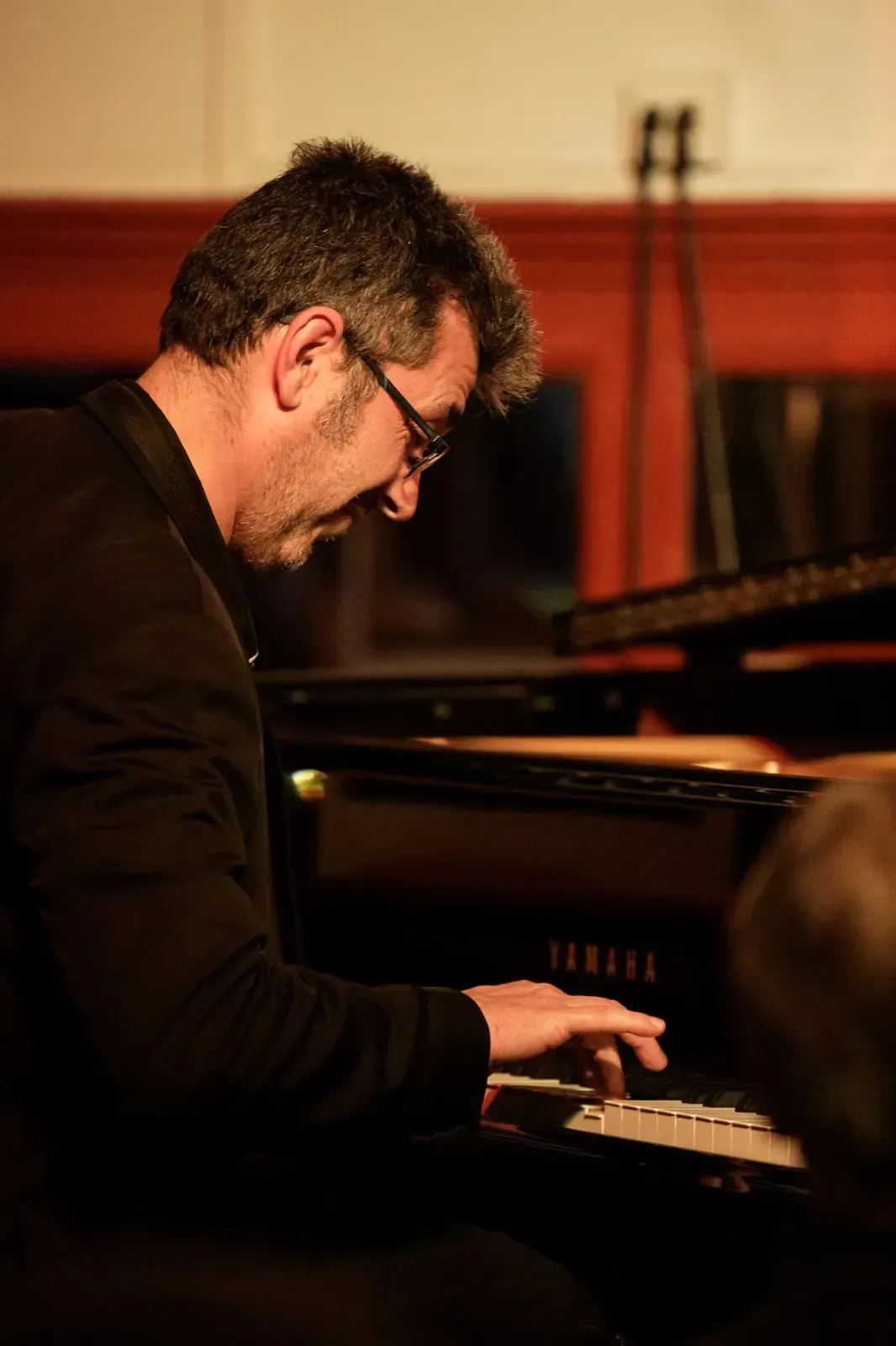 Un hombre toca un piano Yamaha negro en una habitación con poca luz. Lleva gafas y chaqueta negra.