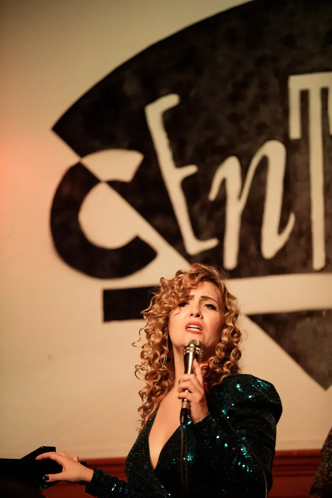 Una mujer canta frente a un micrófono en el escenario, con un cartel de 