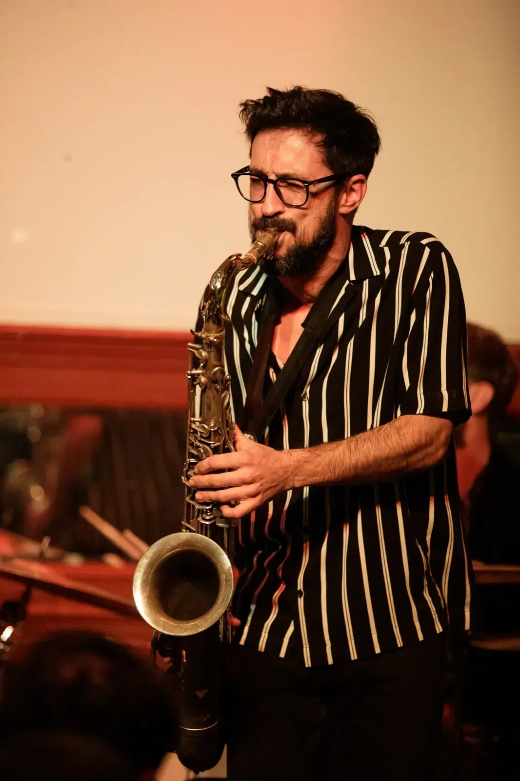 Hombre tocando el saxofón, con gafas y camisa a rayas, actuando en el escenario con una iluminación cálida.