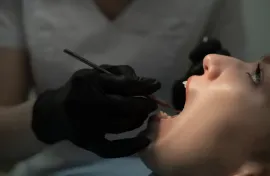Dentista examinando la boca abierta de un paciente con herramientas dentales y guantes negros.