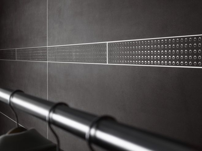 Dunkel geflieste Wand mit einem horizontalen Streifen aus gemustertem Metallprofil über einem silbernen Handlauf