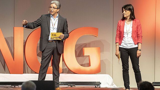 Ein Mann und eine Frau stehen auf einer Bühne vor einem großen orangefarbenen Buchstaben G.