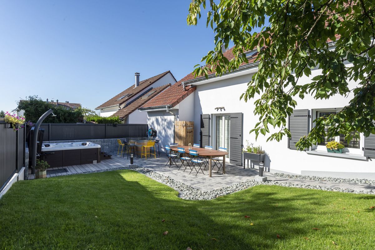 Partie terrasse dans le jardin d'une maison avec table et chaises à proximité d'un spa ouvert.
