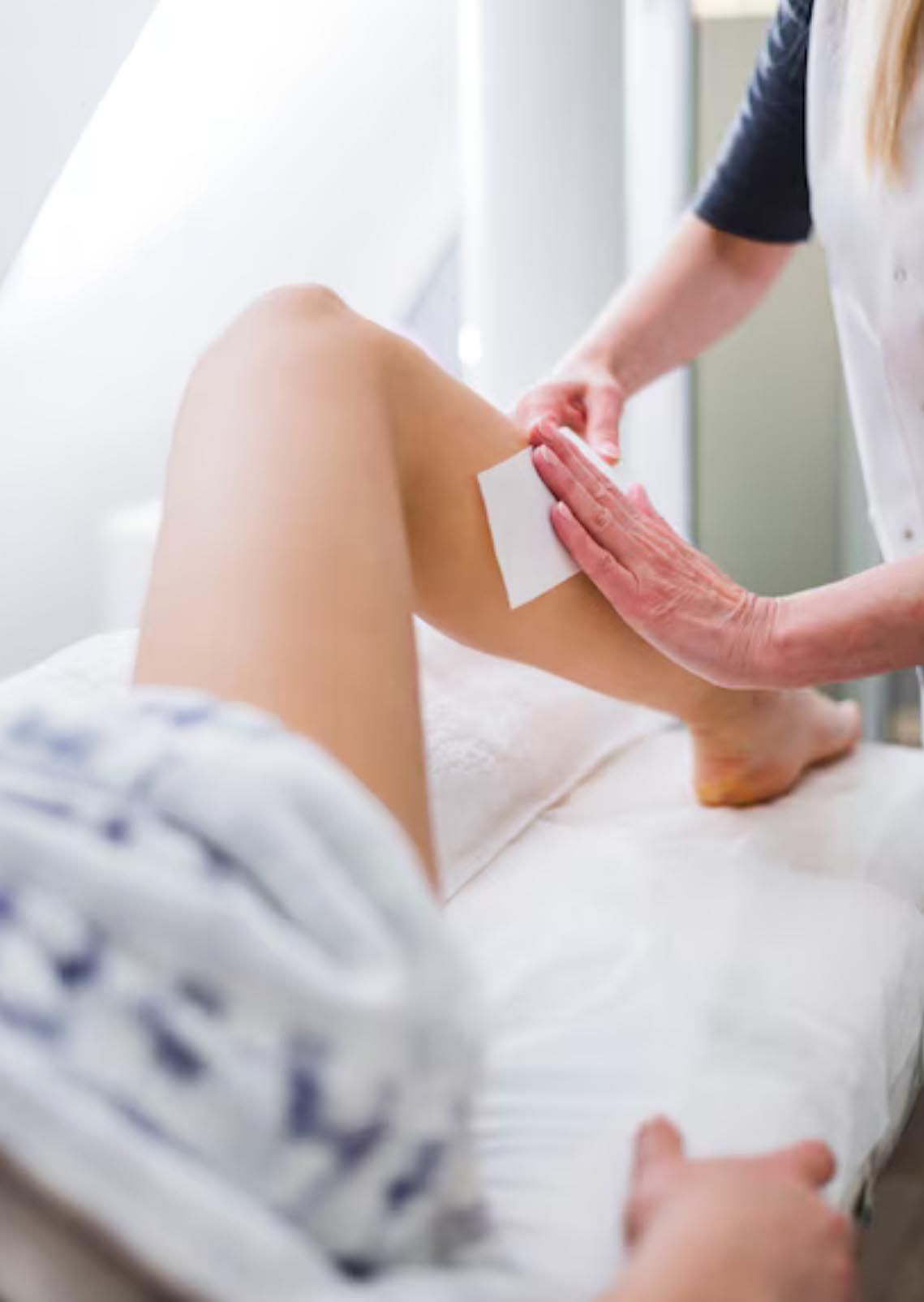 Eine Person lässt sich in einem Salon das Bein wachsen. Jemand hält einen Wachsstreifen an das Bein.