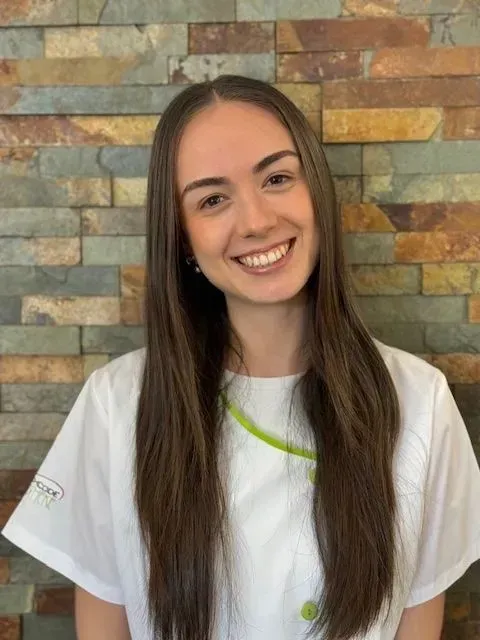 Frau in einem weissen Mantel mit grünem Kragen, lächelnd. Vor einer Backsteinmauer.