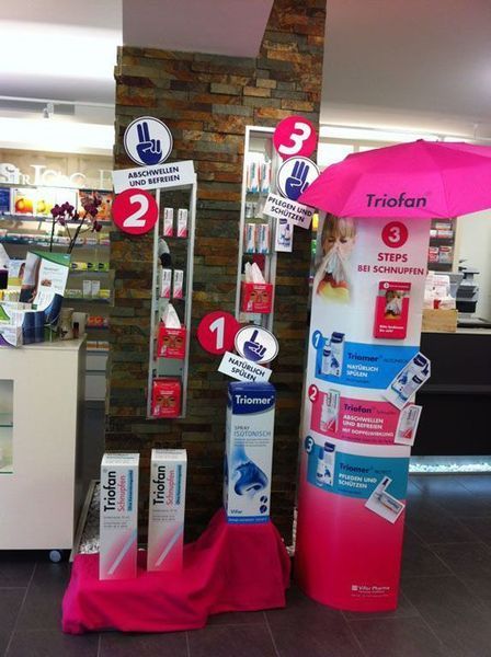 Apothekenauslage: rosa-weisse Trioifan-Produkte, nummerierte Stufen mit Handsymbolen, unter einem rosa Regenschirm.