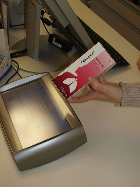 Eine Hand hält eine rosa Medikamentenpackung über einen Scanner an einem Apothekenschalter.
