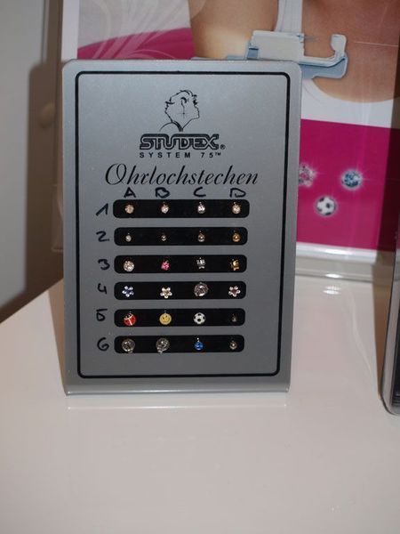 Präsentation von Ohrsteckern zum Ohrlochstechen. Silbernes Schild mit Reihen von Steckern. Schriftzug „System 75“ und „Ohrlochstechen“.