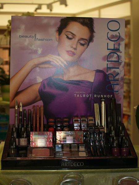 Ein Make-up-Display im Art-Deco-Stil mit einer Frau in einem violetten Kleid auf dem Poster. Die Produkte sind auf einem dunklen Holzständer angeordnet.