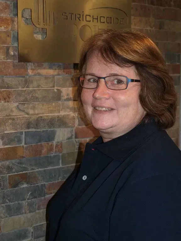 Eine Frau mit langen braunen Haaren lächelt vor einer Backsteinmauer. Sie trägt eine weisse Uniform mit grünem Besatz.