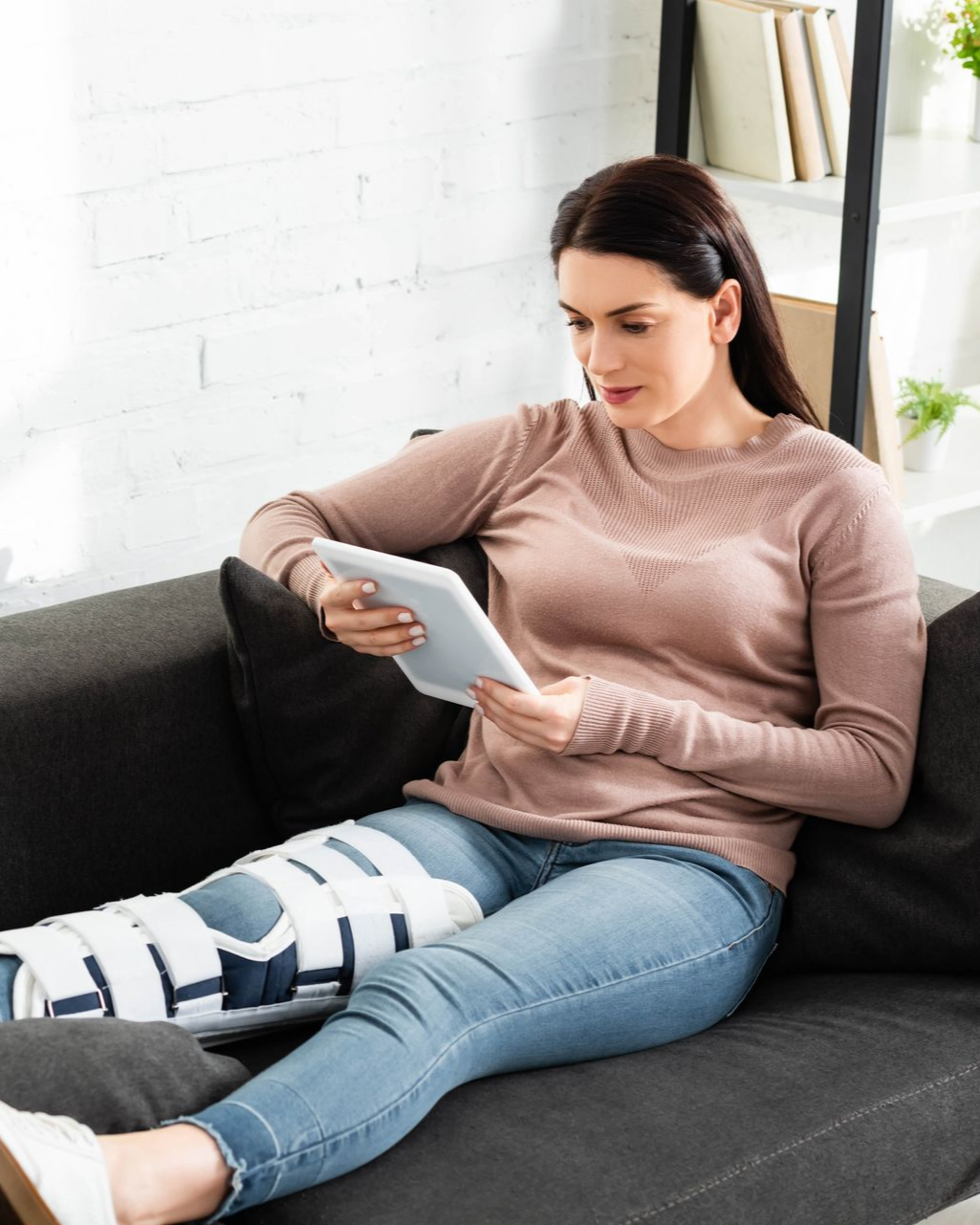 Femme qui regarde un tablette, allongée sur son canapé avec une attelle au genou droit