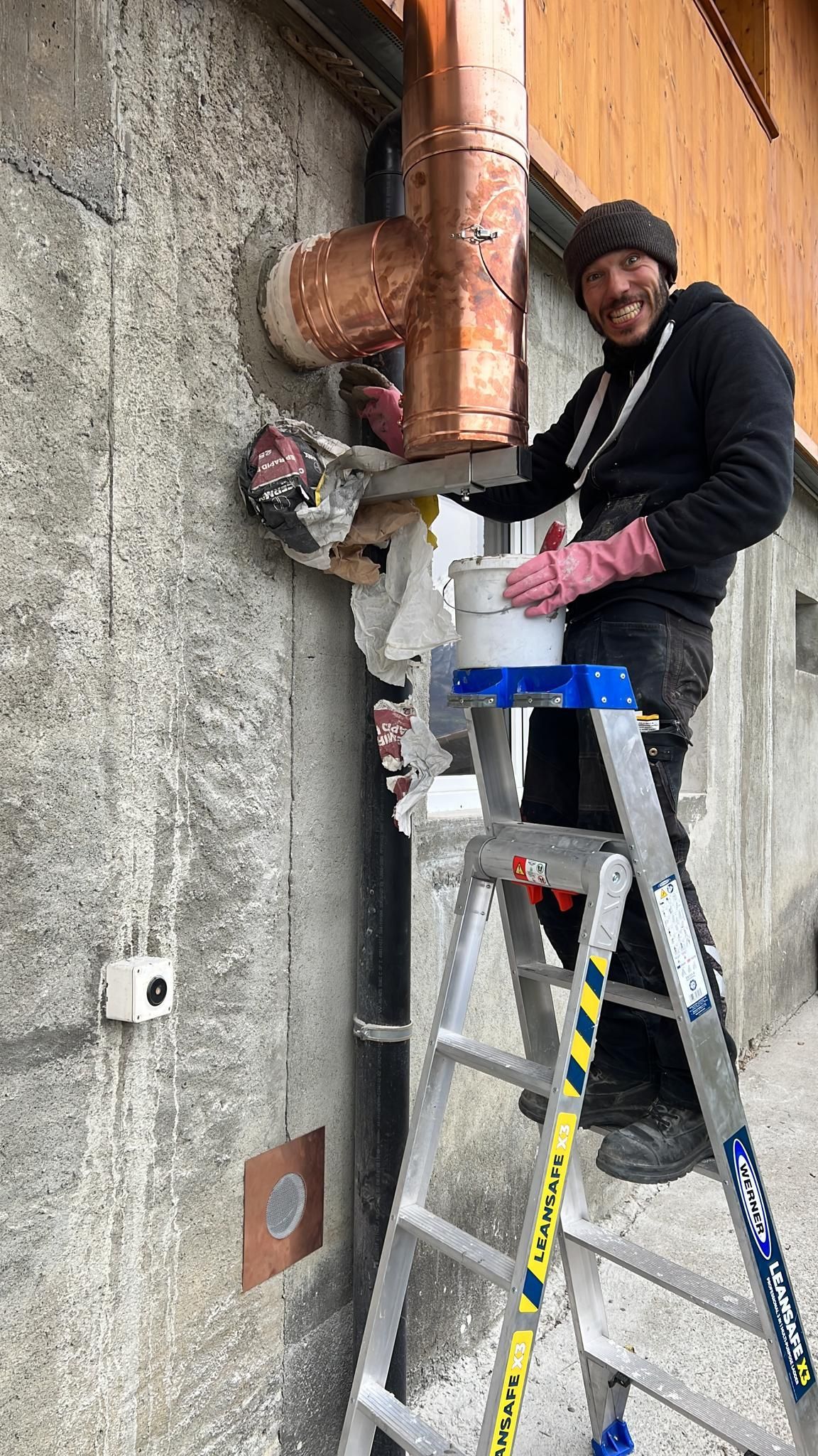 Un homme sur une échelle installe un tuyau en cuivre sur un mur en béton, portant des gants, un chapeau et souriant.