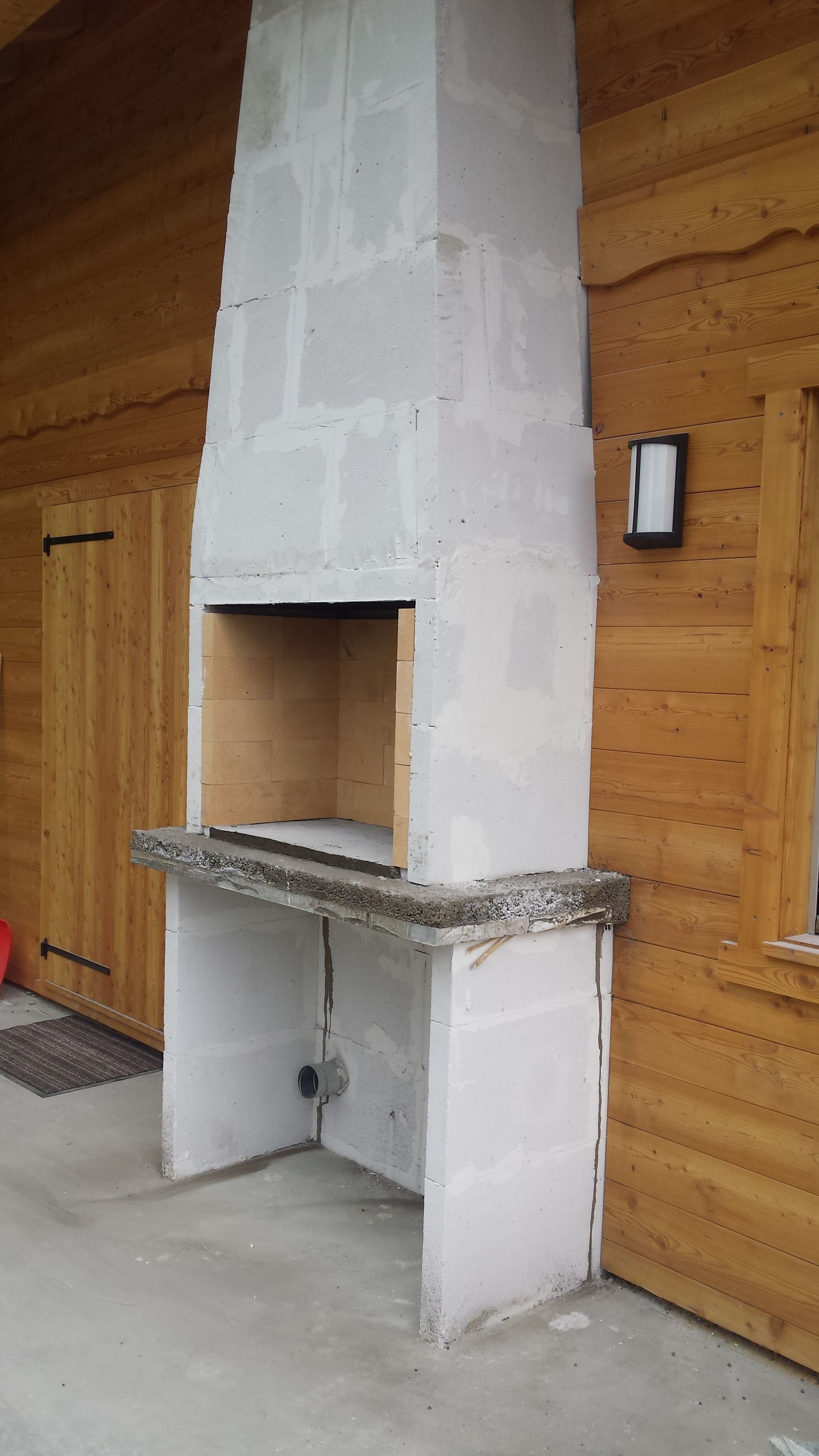 Cheminée extérieure brute contre un revêtement en bois. Le foyer est visible et une petite lumière est fixée au mur.