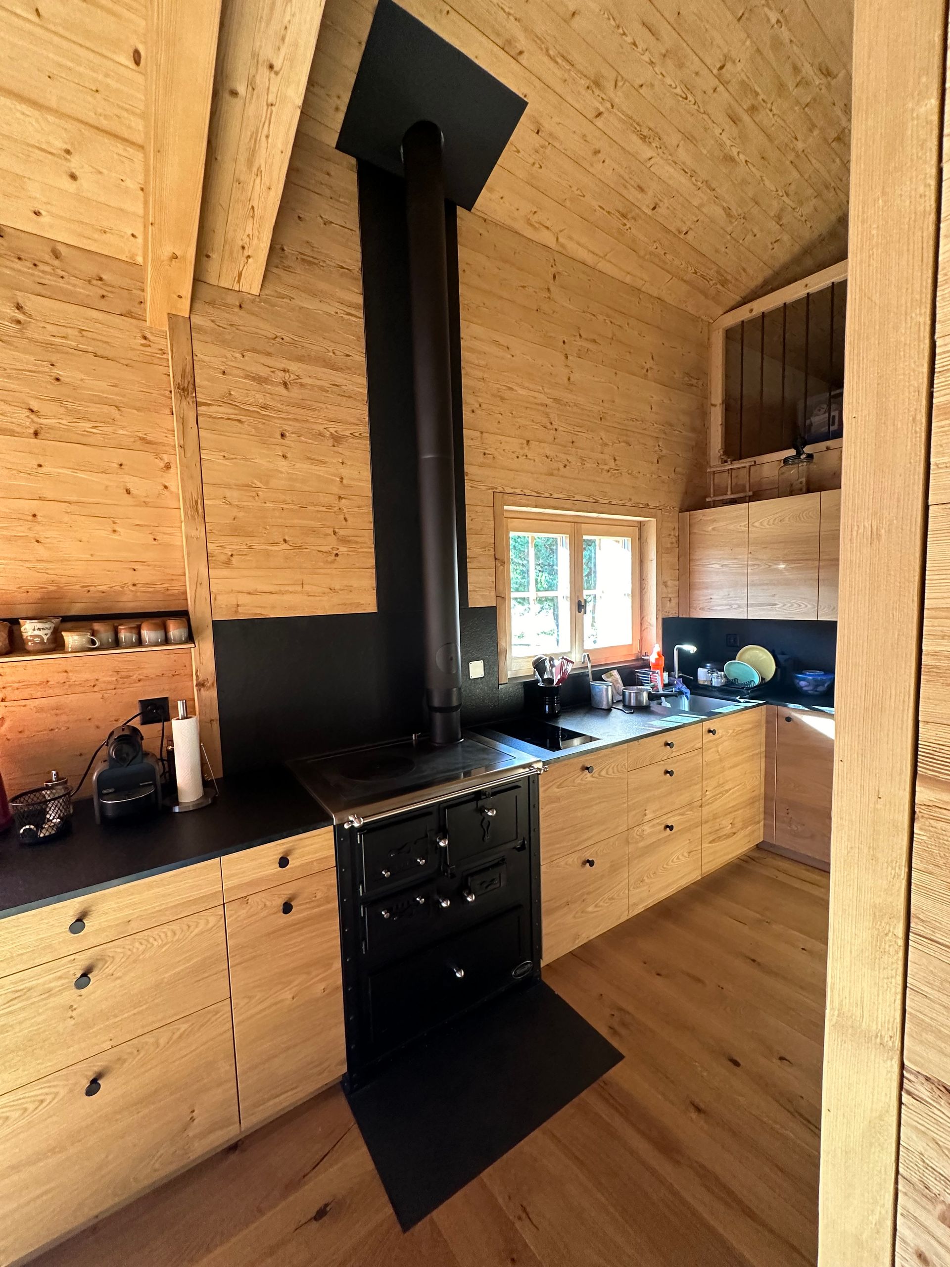 Une cuisine rustique en bois avec un poêle et une cheminée noirs, des armoires en bois et une fenêtre.