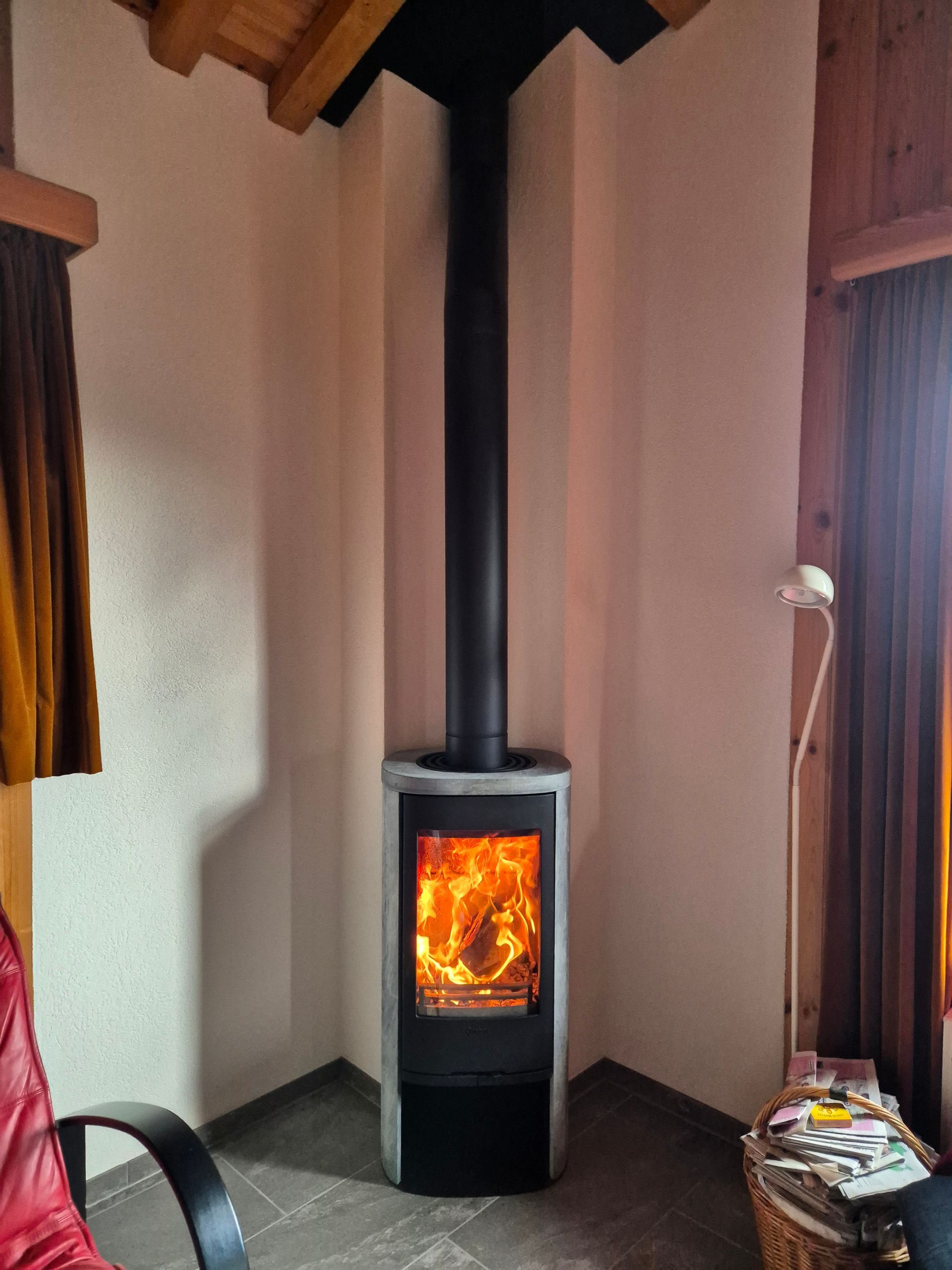 Un poêle à bois trône dans un coin d'une pièce, avec des flammes visibles à l'intérieur. Une cheminée noire s'élève du poêle.