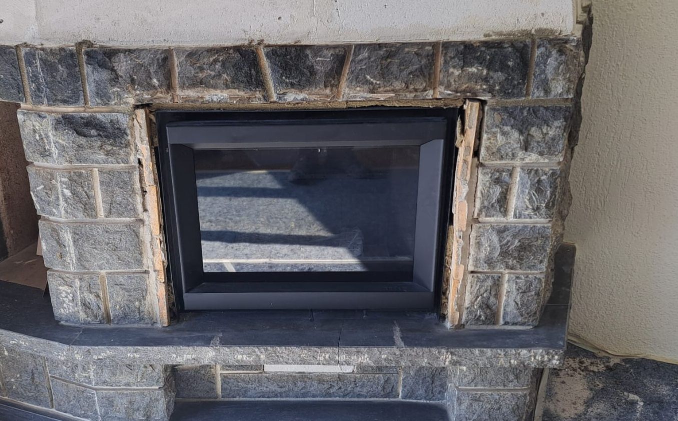 Cheminée avec encadrement en pierre gris foncé, cadre noir et porte vitrée. Le décor semble être extérieur.