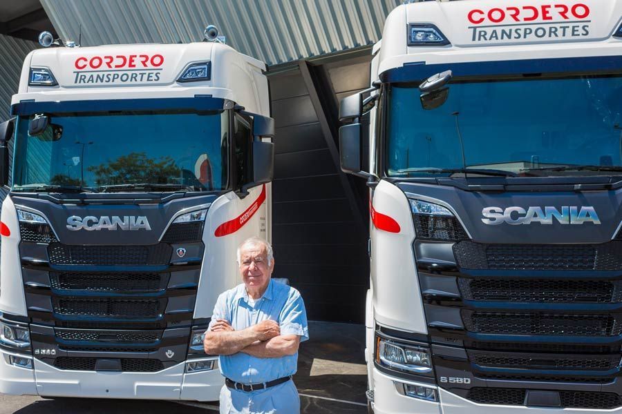 Una persona permanece de pie con los brazos cruzados entre dos camiones Scania blancos con la marca "Cordero Transportes" en unas instalaciones.