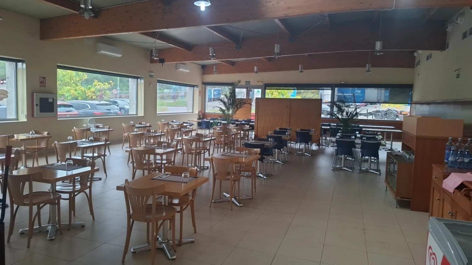 Cafetería vacía con mesas y sillas de madera, grandes ventanales y suelo de baldosas.