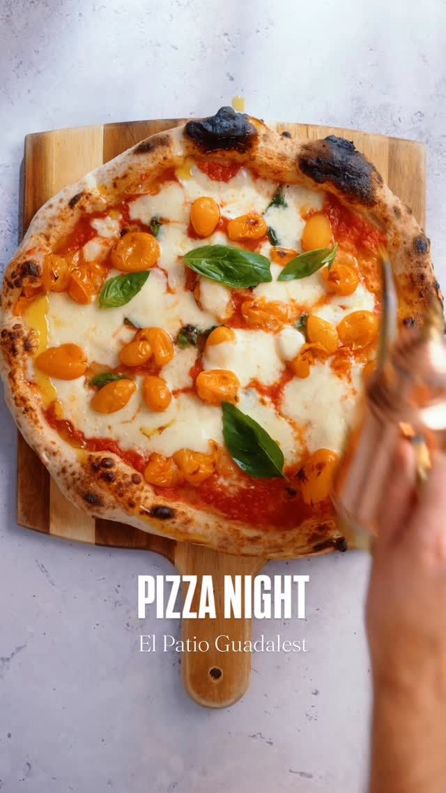 Pizza fresca con albahaca sobre una tabla de madera, con una mano sosteniendo una porción; el texto dice