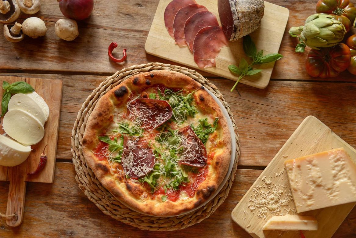 Pizza sobre una mesa de madera con lonchas de embutido, queso, ajo y hierbas aromáticas cerca.