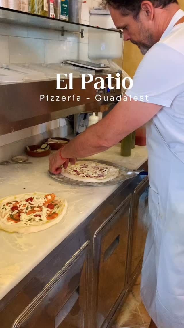 Chef colocando pizzas en el mostrador de la cocina de una pizzería con letrero de “El Patio”.