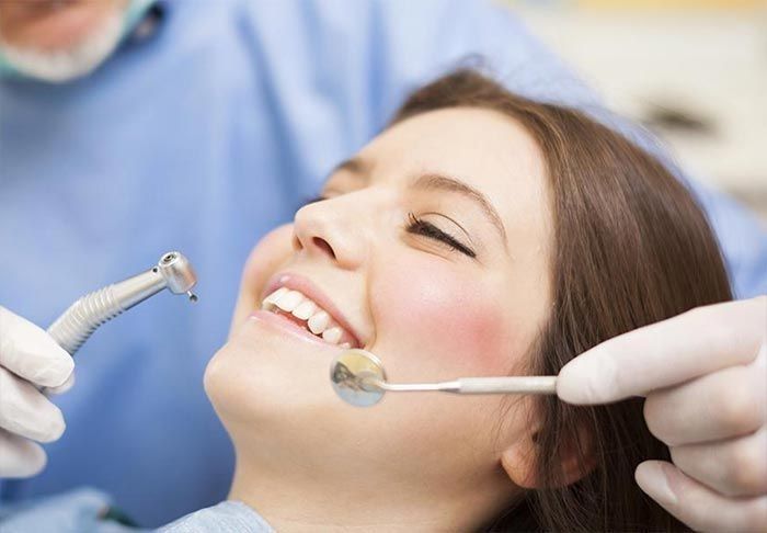 Mujer en una cita con el dentista, sonriendo con la boca abierta; dentista sosteniendo herramientas.