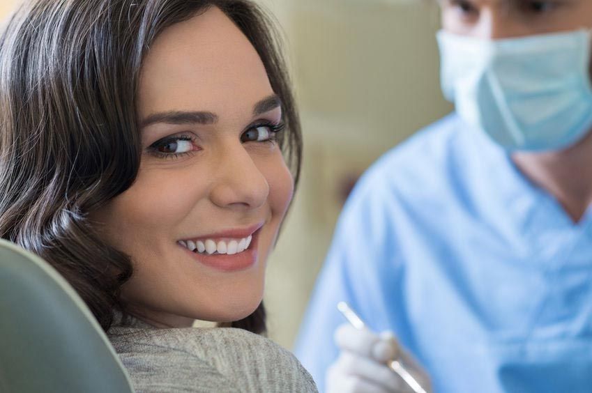 Mujer sonriendo en una silla dental; dentista con uniforme azul y mascarilla.