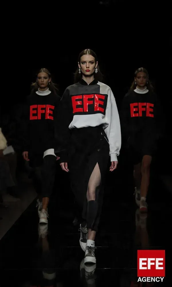 Tres modelos desfilando en una pasarela frente a un telón de fondo rojo con la inscripción