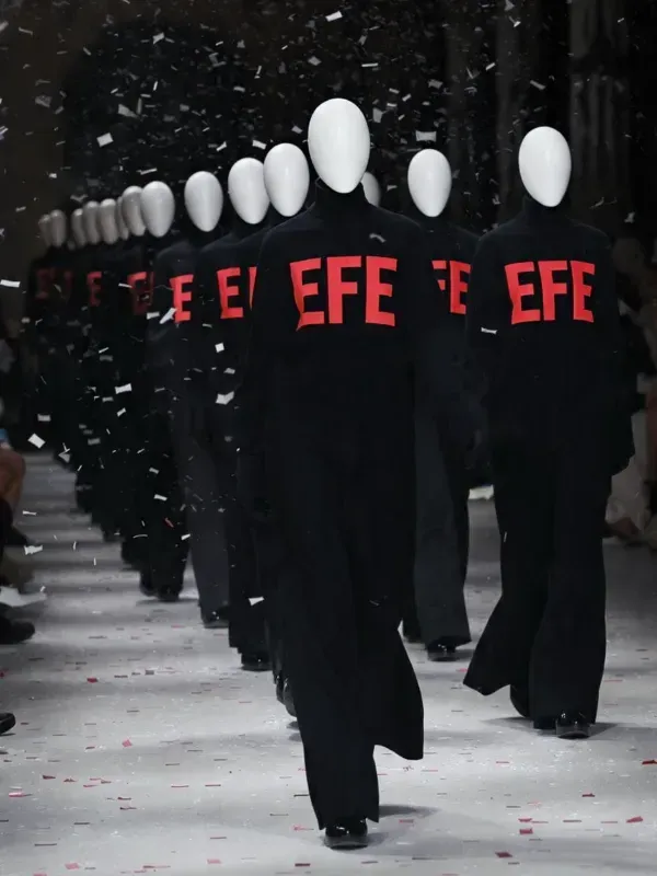 Fila de artistas enmascarados con túnicas negras y letras rojas de EFE marchando entre confeti que cae