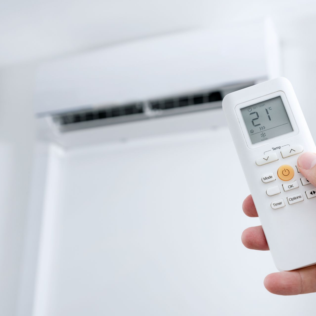 Une main tenant une télécommande blanche est pointée vers un climatiseur mural. L'écran affiche « 21 ».