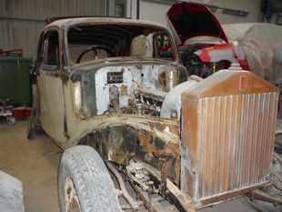 Carrocería vieja y oxidada en un garaje, con la parte delantera al descubierto, una gran parrilla y un segundo coche rojo detrás.